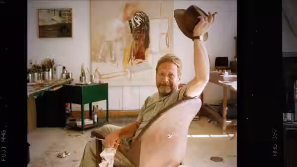Self (Martin Mull) raises a hat while seated in a kitchen