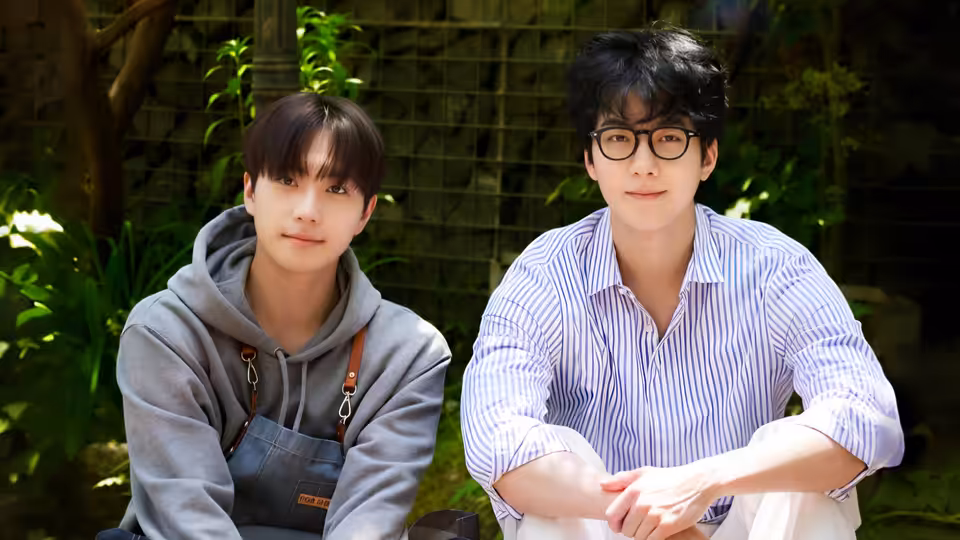 Two young men sit side by side in a leafy courtyard