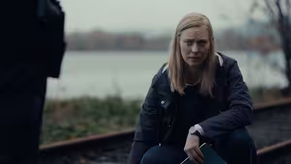 Meg (Deborah Ann Woll) sits beside the water, looking ahead with a tense expression