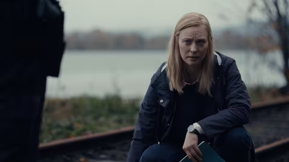 Meg (Deborah Ann Woll) sits beside the water, looking ahead with a tense expression