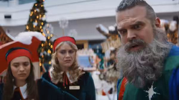 Santa Claus (David Harbour) stands with two elf workers in a decorated room