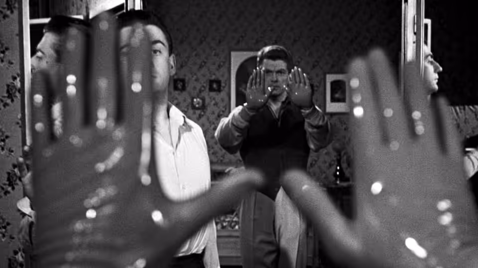 A man raises his hands before a mirror as others watch from the room