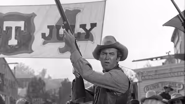 A cowboy raises his rifle beneath a large town sign