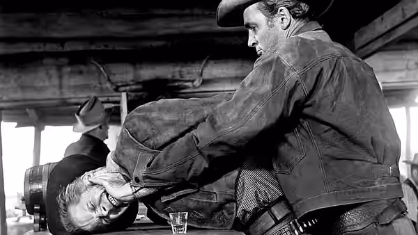 A standing cowboy pins a prone man across a table inside a wooden building