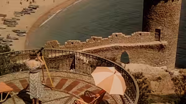 A figure holding a parasol stands on a seaside terrace above moored boats