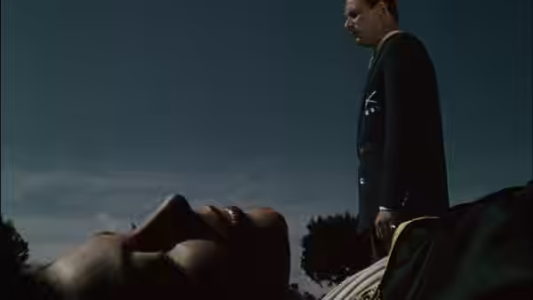 A suited man stands over a reclining figure on the beach