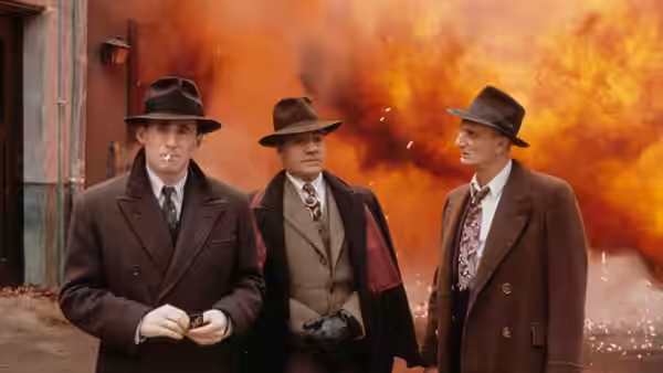 Three men in hats and coats standing before a fiery explosion