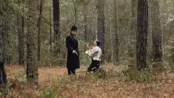 A man pointing a gun at another kneeling man in a forest