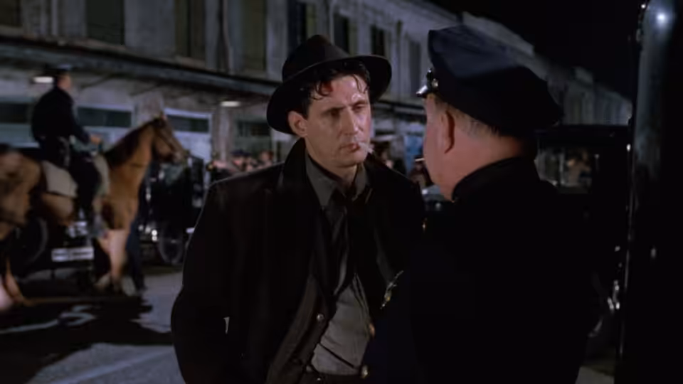 Tom Reagan (Gabriel Byrne) speaking with a police officer on a city street at night