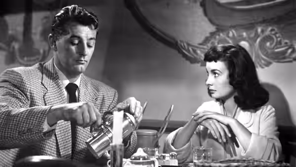 Frank Jessup (Robert Mitchum) pours a drink at the table as Diane Tremayne (Jean Simmons) watches
