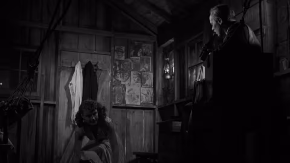 A man crouches in a dim room as another watches from the raised platform