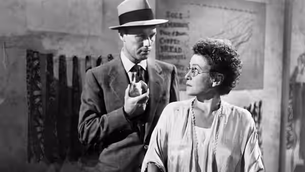 A suited man in a fedora speaks with a bespectacled woman indoors