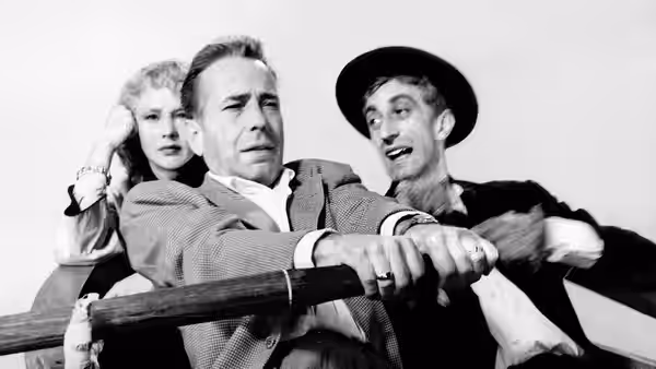 Billy Dannreuther (Humphrey Bogart) grips a railing as a woman and a man in a hat crowd behind him