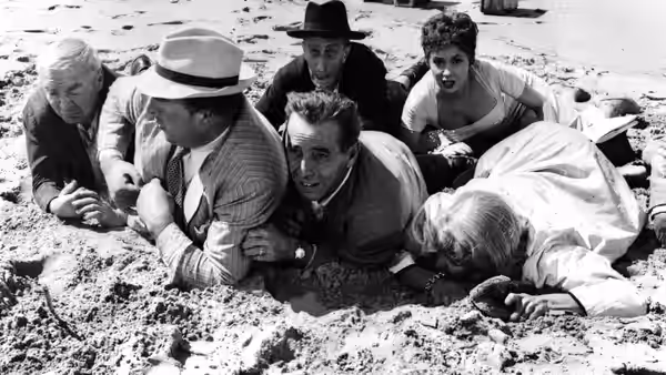 A group of travelers crouch together on a sandy beach beside the water