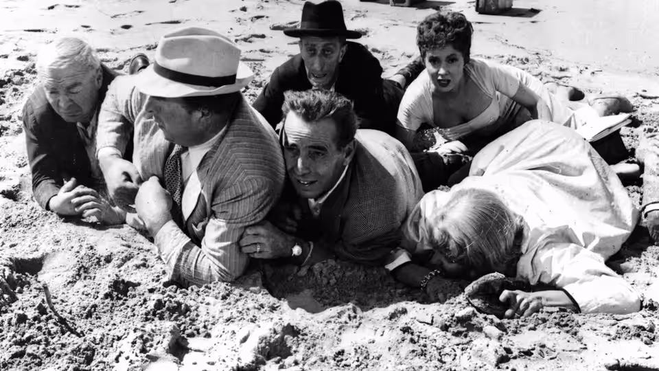 A group of travelers crouch together on a sandy beach beside the water