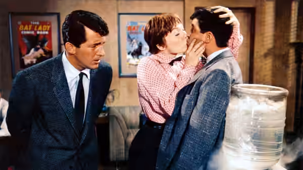 A woman kisses a suited man while another man watches in a room lined with posters