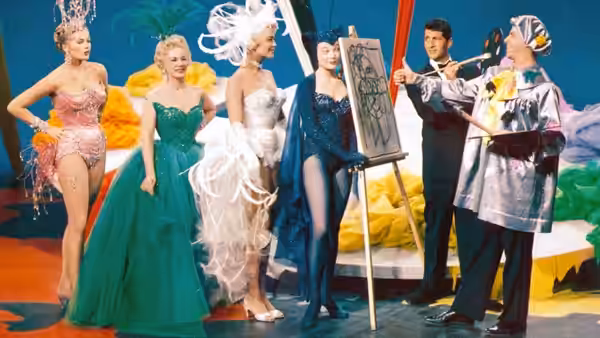 A man presents a drawing board to fashion models posing in a colorful studio