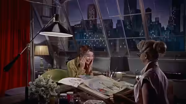 A woman at a drafting table speaks with another woman in a high-rise office