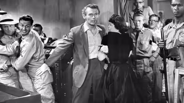A man and woman stand together as a crowd of men surrounds them