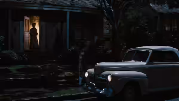 A silhouetted woman stands in a lit doorway as two cars are parked outside the house