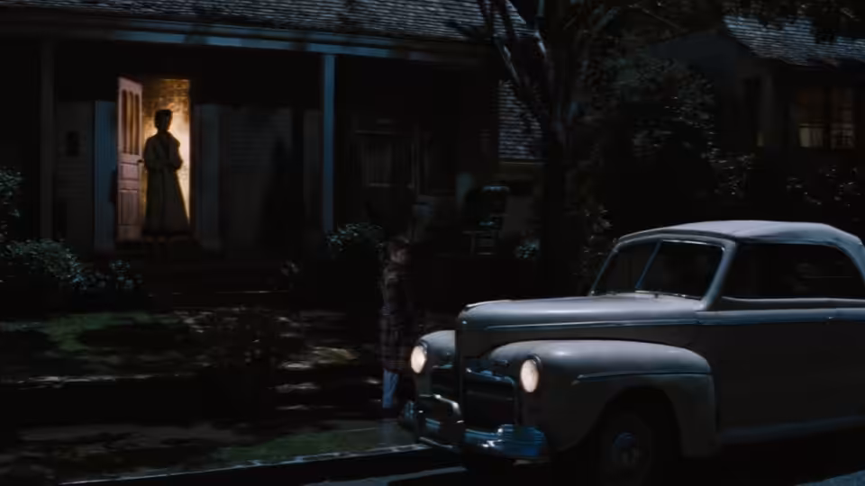 A silhouetted woman stands in a lit doorway as two cars are parked outside the house