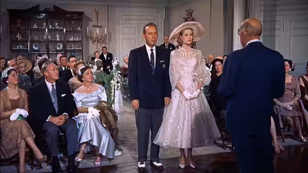 Tracy Lord (Grace Kelly) stands beside George Kittredge (John Lund) before seated wedding guests