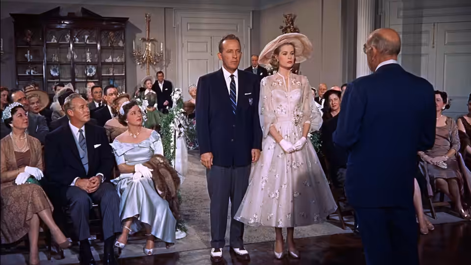 Tracy Lord (Grace Kelly) stands beside George Kittredge (John Lund) before seated wedding guests