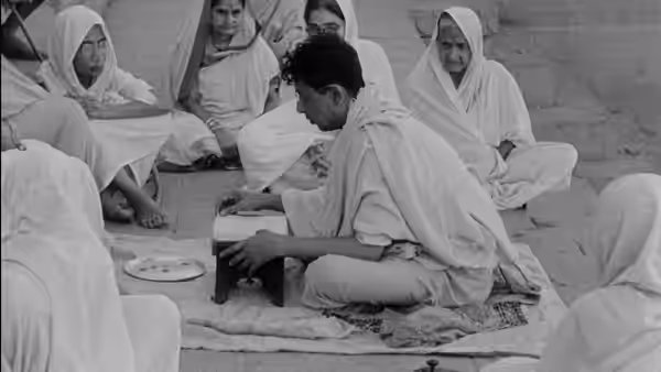 Teenage Apu Roy sits among women in white saris, working at a low desk