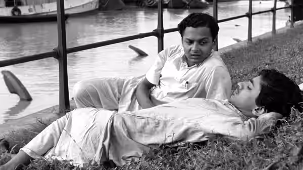 A man sits beside teenage Apu Roy as he lies on a grassy riverbank