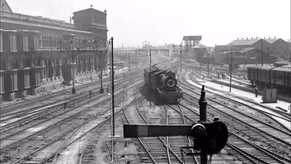 A steam locomotive approaches along railway tracks beside a station