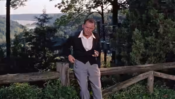 Dave Hirsh (Frank Sinatra) leans against a wooden fence overlooking a wooded lakeside