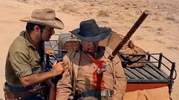 A cowboy holds a seated man beside a wagon in the desert