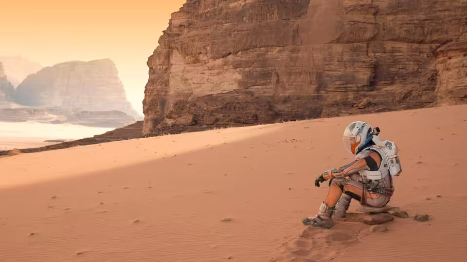 An astronaut in an orange spacesuit sits on a rock in a sandy desert