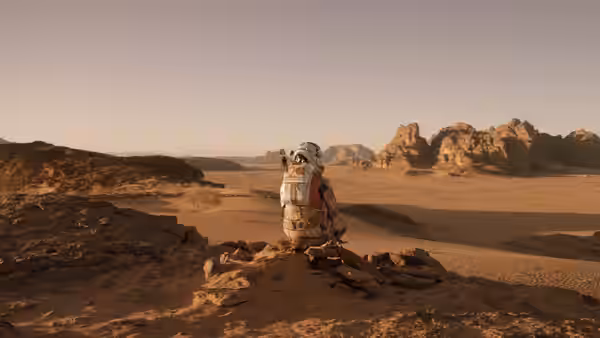 An astronaut stands on a rocky ridge overlooking a vast sandy desert landscape