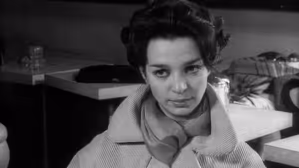 Lelia (Lelia Goldoni) sits among tables wearing a coat and scarf