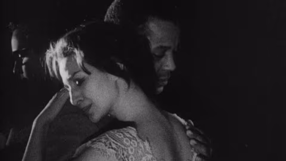 A woman leans forward while a man stands close behind her in the dark