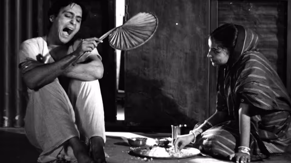 A man playfully holds a fan while a woman serves food at a low table