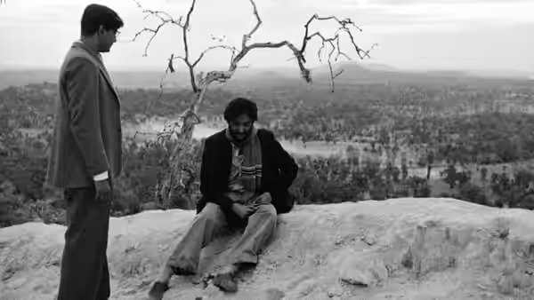 Two men meet on a rocky overlook above a wooded landscape