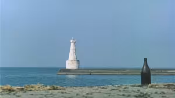 A lighthouse stands offshore as a bottle rests by the sea