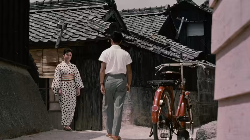 A woman in a patterned kimono faces a man beside a rickshaw outside a tiled-roof house