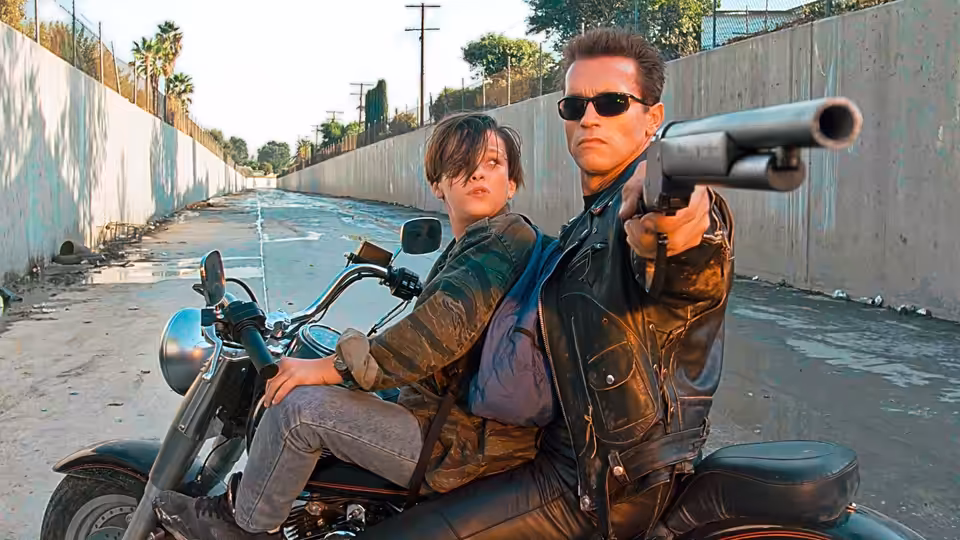 John Connor (Edward Furlong) rides a motorcycle as The Terminator (Arnold Schwarzenegger) aims a shotgun