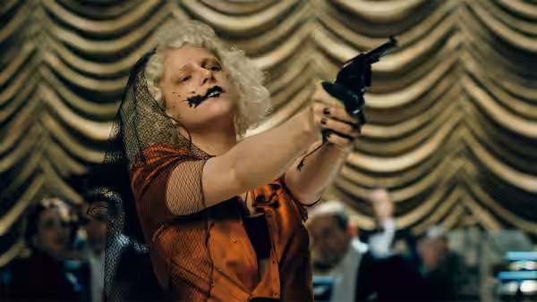 Woman in orange dress and veil aims a revolver upward in a theater