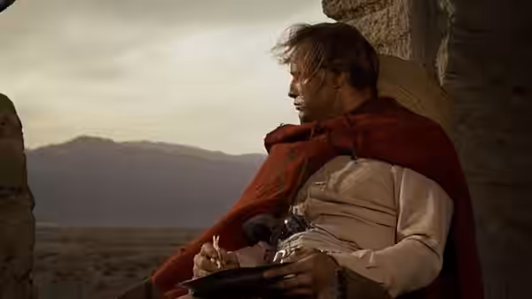 A man wrapped in a red blanket sits beside a window overlooking the landscape