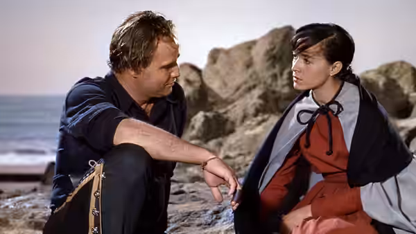 A man and woman sit facing each other on a rocky shore