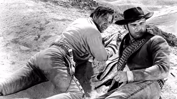 A man crouches beside a seated cowboy holding a rifle on a rocky hillside