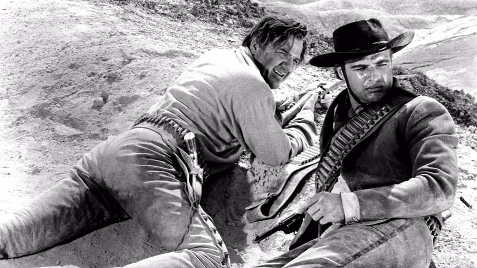 A man crouches beside a seated cowboy holding a rifle on a rocky hillside