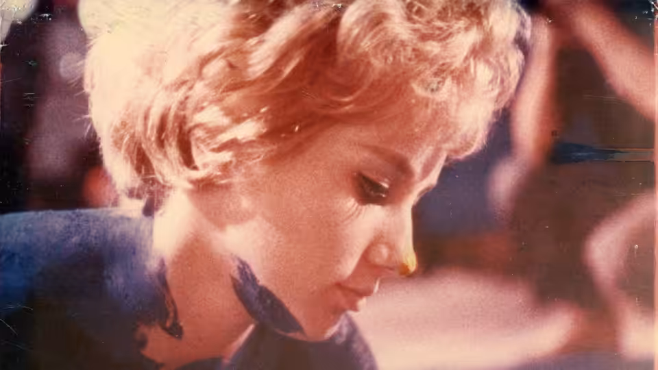 A blonde woman in a blue outfit looks down against a dark background