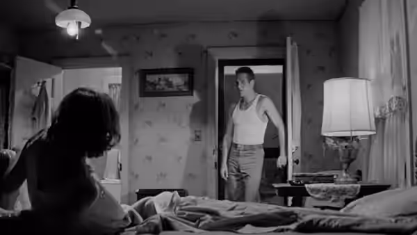A woman sits beside a bed as a young man stands in the doorway