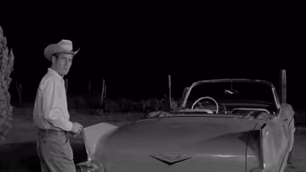 A cowboy stands beside a parked car at night