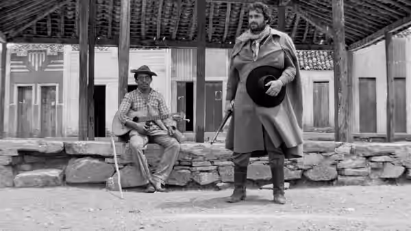 Corisco (Othon Bastos) stands holding his hat beside a seated man on a village porch
